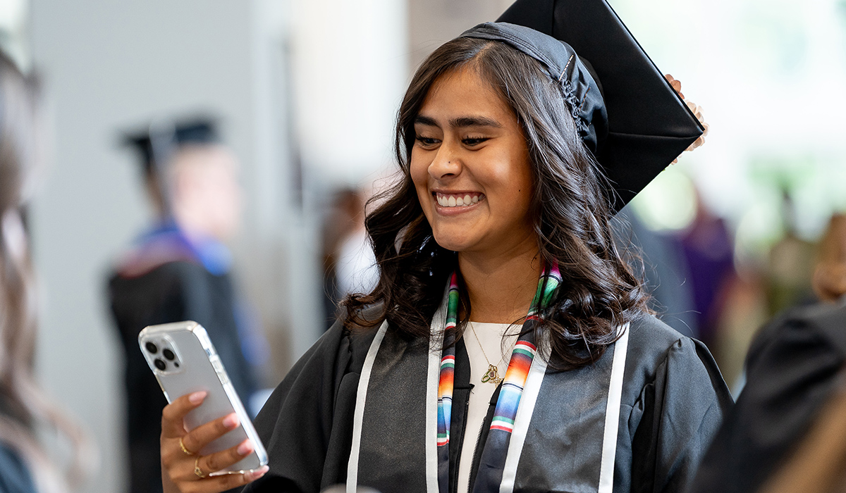 A graduate wearing a cap and gown smiles down at her phone.