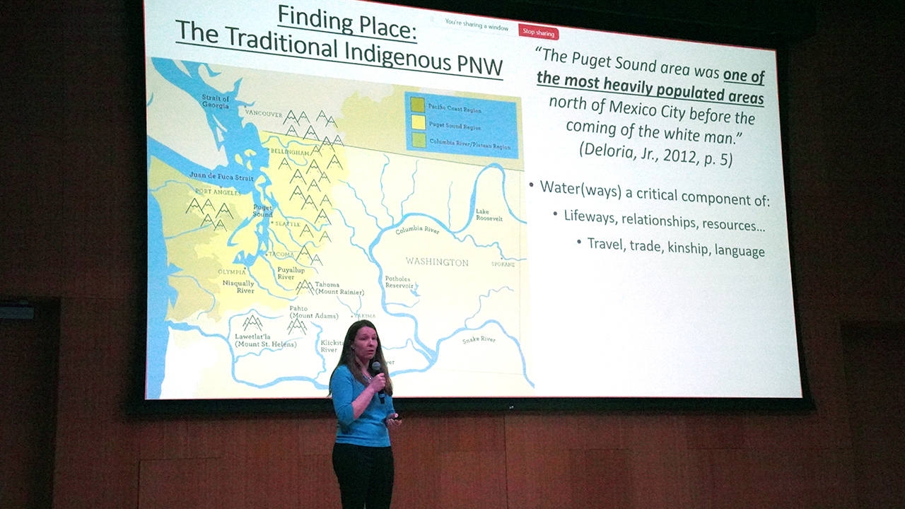 Professor Kestrel Smith stands in front of a presentation showing a map where Indigenous people lived in the Pacific Northwest.