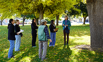 Dr. Joan Qazi stands beneath a tree with a group of students during an Arbor Day activity.
