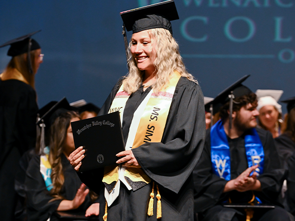A WVC Omak graduate wearing a gold stole walks across the stage holding her diploma.